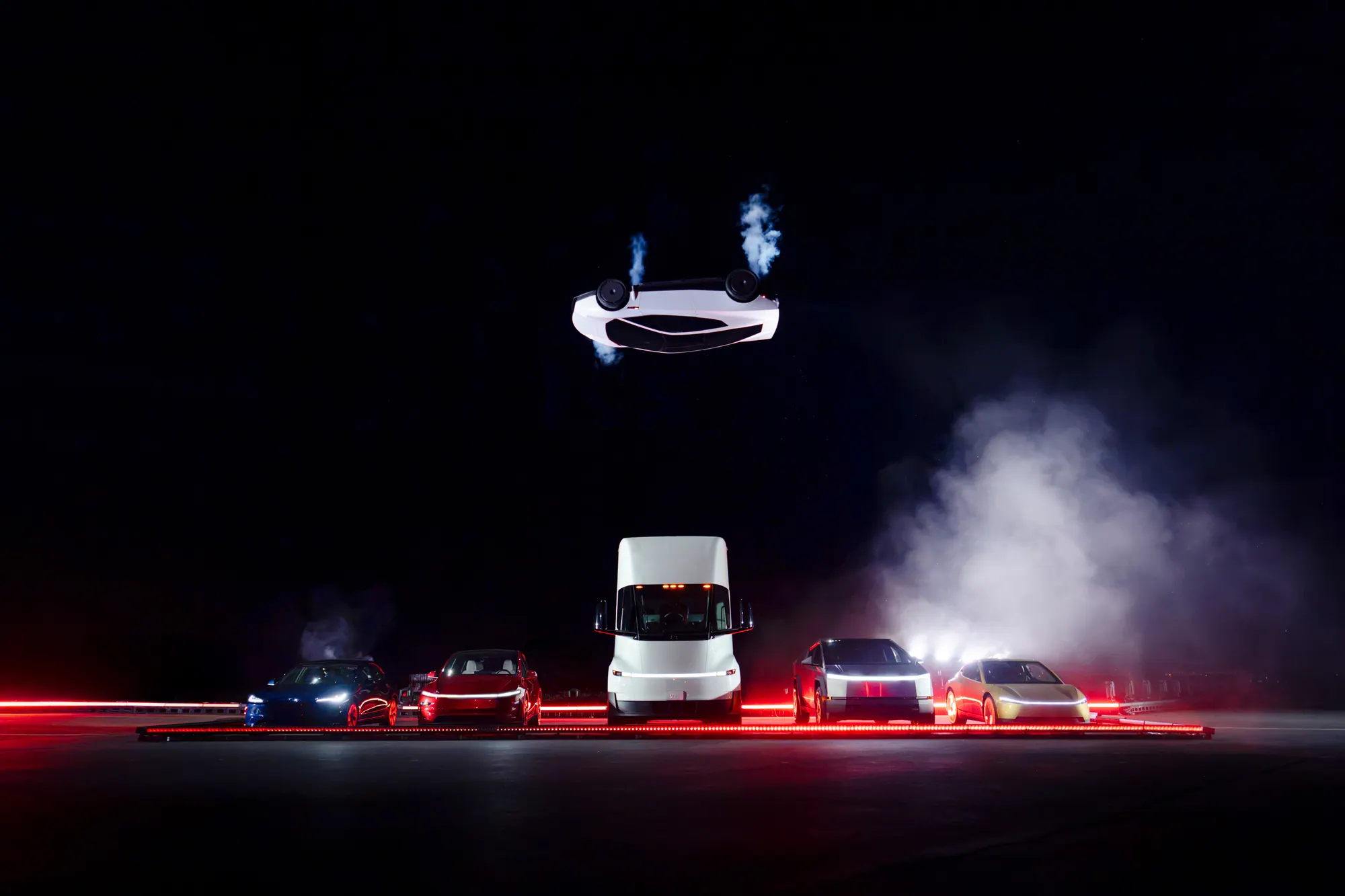 The white Roadster fully inverted, wheels toward the sky, above a Semi and other Tesla vehicles. A red light strip runs along the ground.