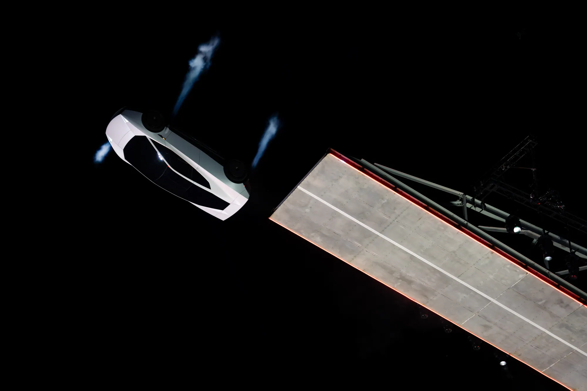 Overhead view of the white Roadster inverted just beyond the end of a ramp, with blue vapour trailing from the car.