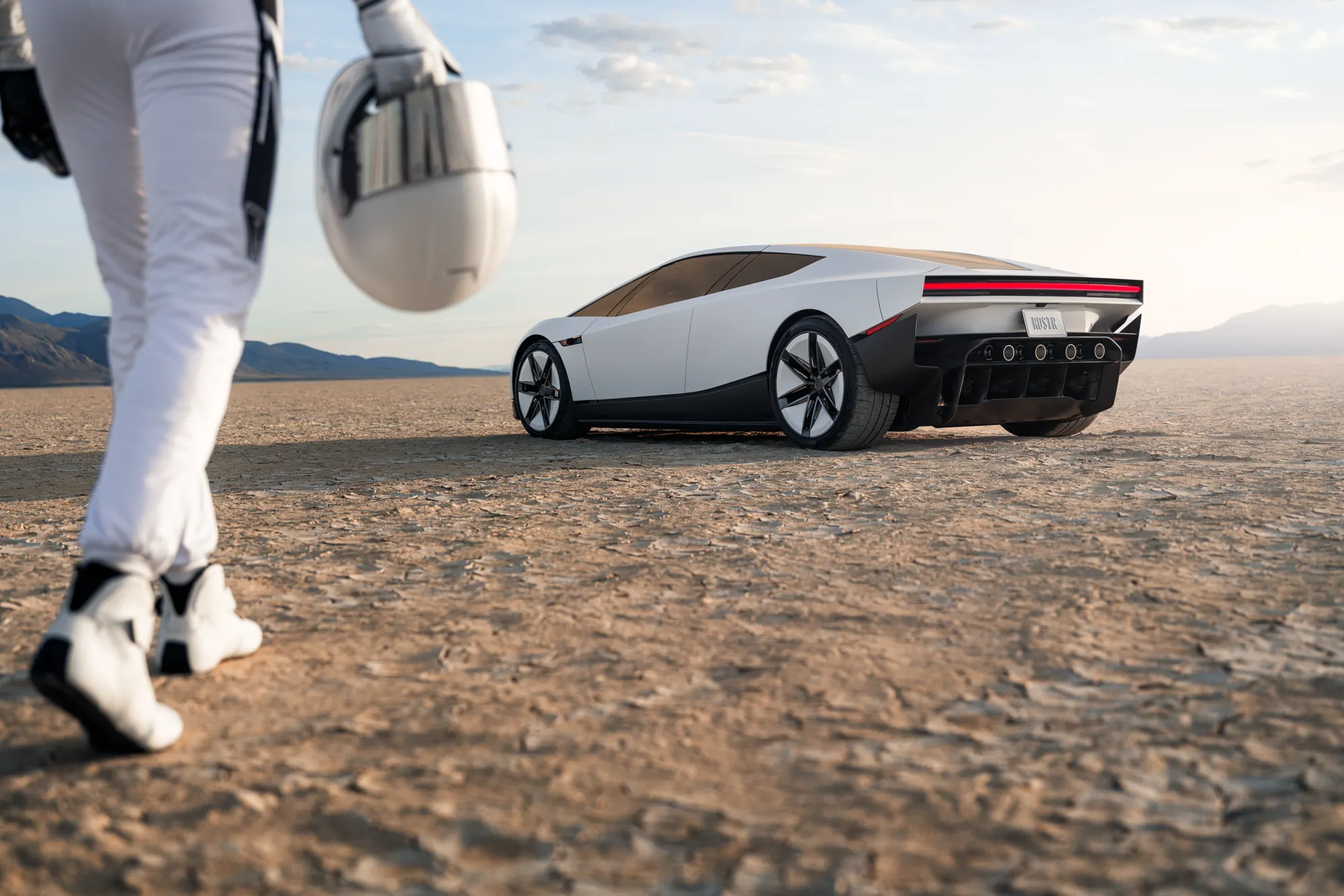 Rear three-quarter view of the white Roadster on a dry lakebed, with a person in a flight suit and helmet in the foreground.