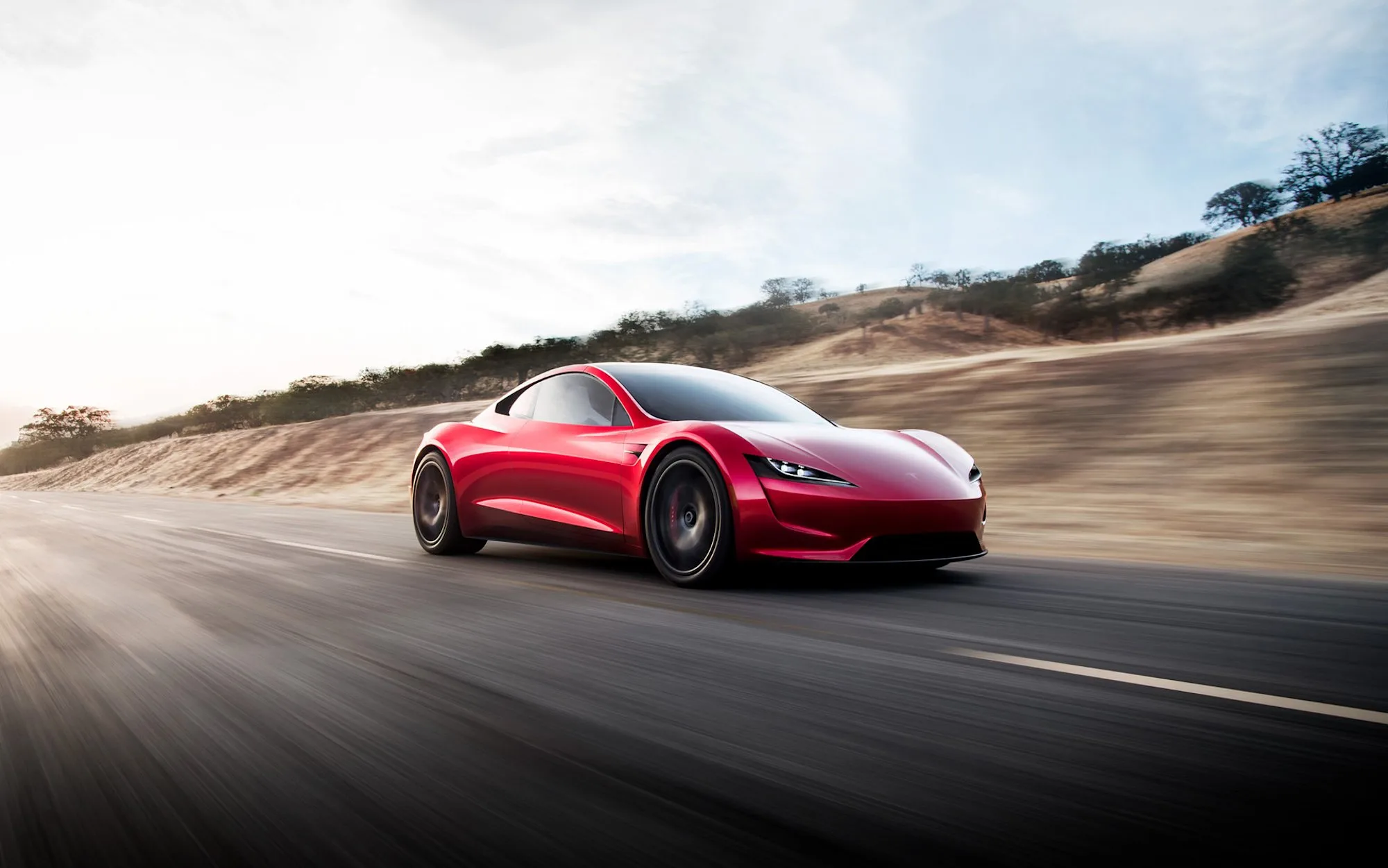 Red 2017 Tesla Roadster prototype driving on a road, with a low nose and conventional headlights. This is the earlier reveal car, not the 2026 design.