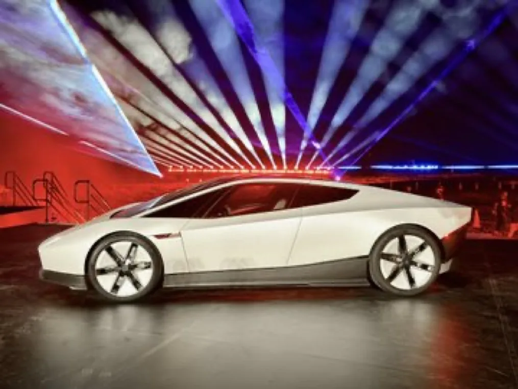White second-generation Tesla Roadster in side profile on a dark stage, with red and blue light beams behind it.